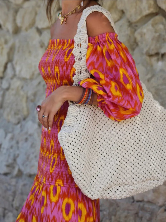 Person wearing a colorful dress holding a white woven bag against a stone wall.