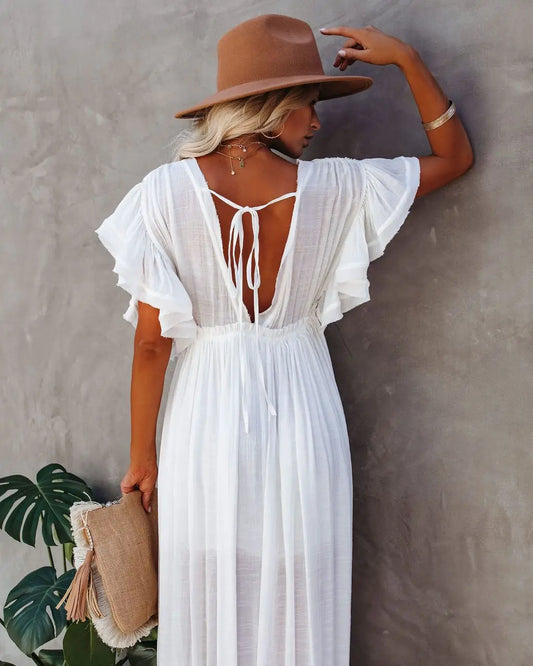 Woman wearing a white dress with a backless design and a brown hat, standing against a plain wall.