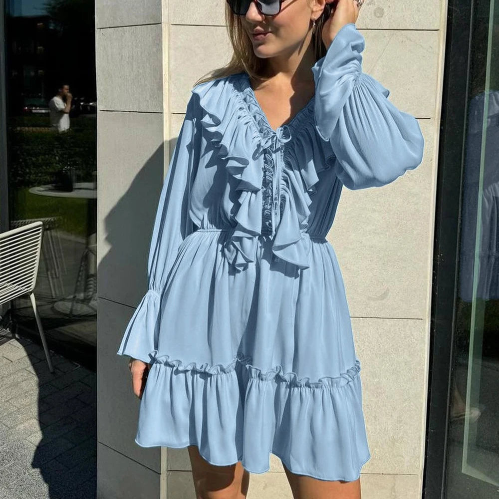 Woman wearing a light blue dress with long sleeves and ruffled layers, standing against a building.