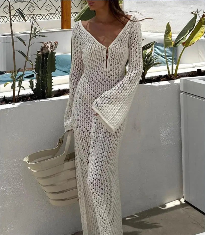 Person wearing a long, light-colored knitted dress standing outdoors with plants and a pool in the background.